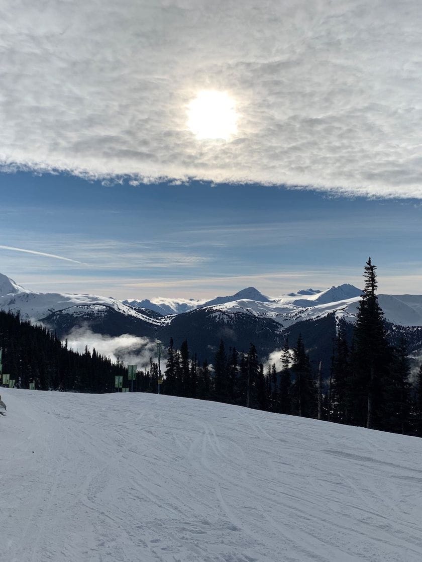 The top of a snowy ski hill with mountain ranges in the distance.