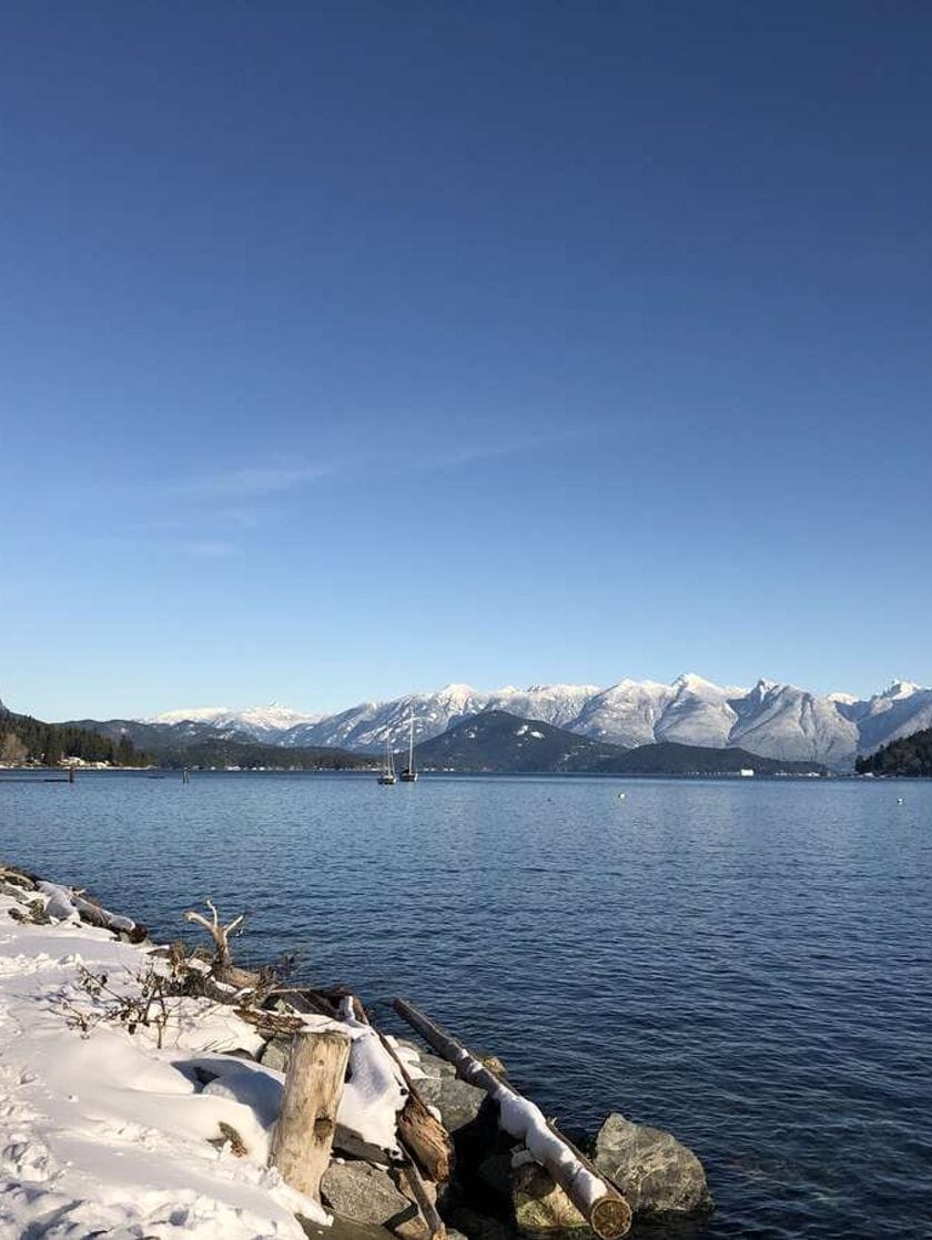 A snowy path alongside a dark body of water where sailboats linger, with snow-capped mountains on the horizon. 