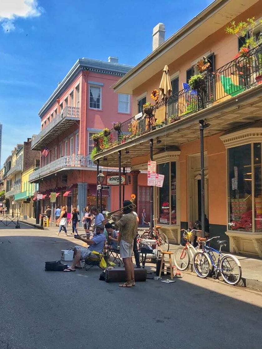 A street flanked by pink and orange two-storey buildings with flowered balconies. In the road, people are walking, bicycles lean against an iron pillar and buskers are performing.