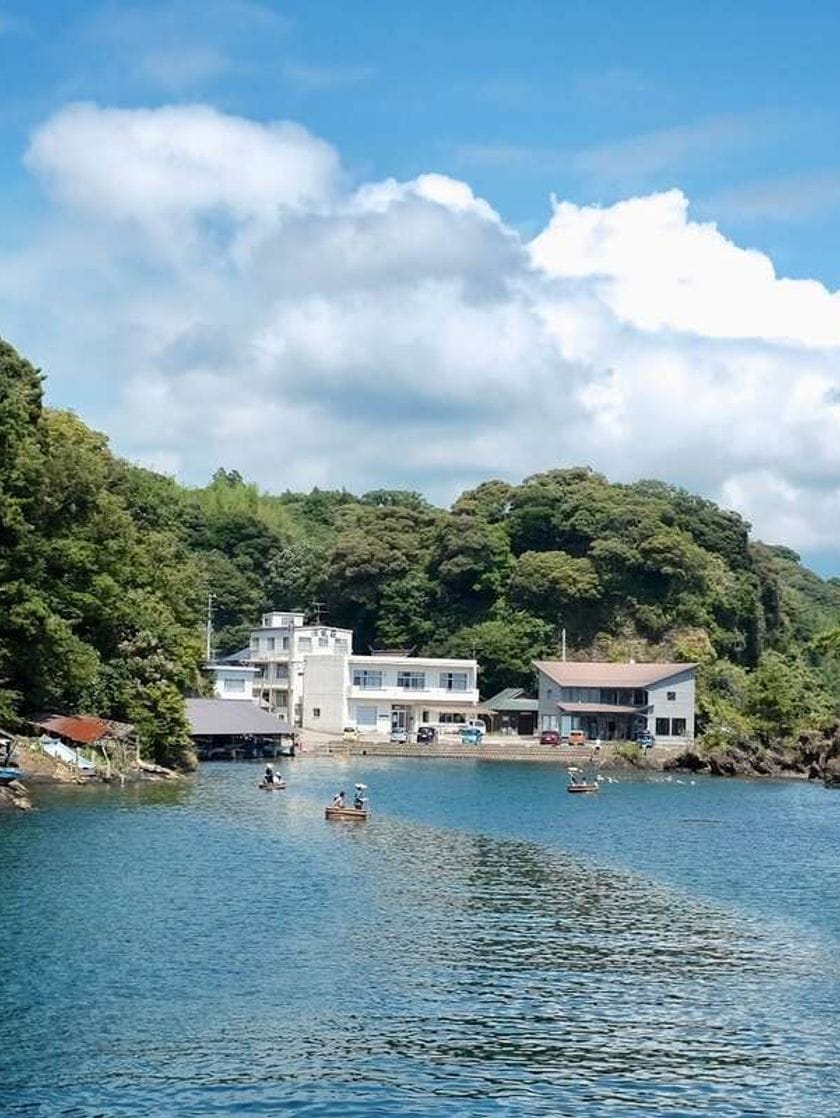 Small groups of people float on a lake in wooden boats. The landscape around the lake is lush and green.