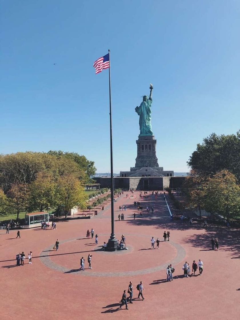 Crowds milling around an American flab on the grounds in front of the Statue of Liberty.