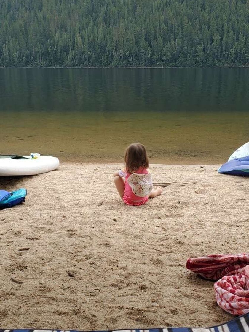 A young girl wearing a pink t-shirt and shorts sits on a sandy beach, facing a lake with pine trees visible on opposing shore.