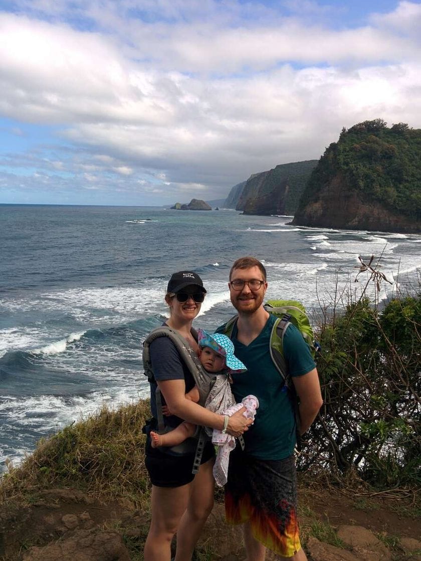 A man and woman stand next to each other in front of an ocean shore with rocky cliffs in the distance. The woman is carrying a baby in a harness.