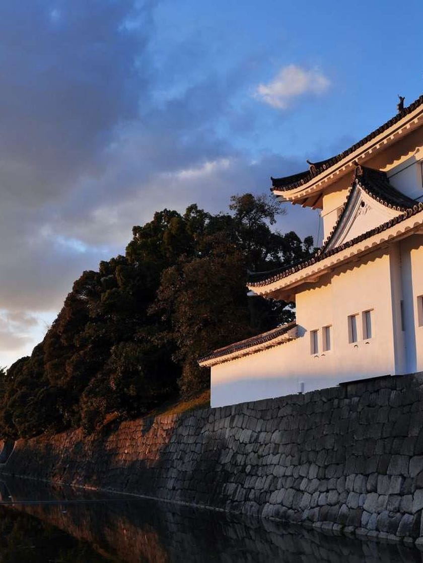 The sun sets, casting soft light on the side of a white building at the edge of a river.