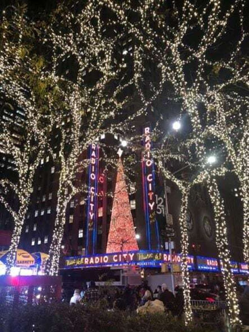 The neon Radio City Music Hall sign surrounded by trees lit up with white Christmas lights at night.