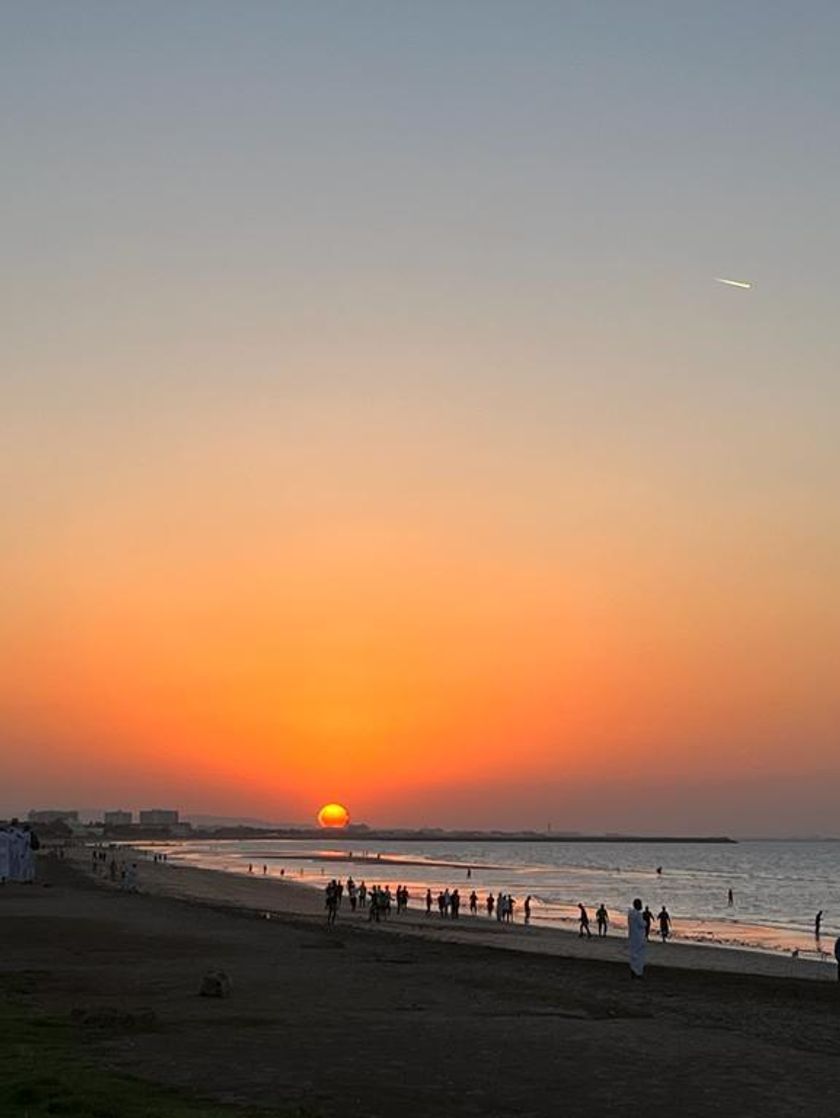 A sun sets in a deep orange sky over the ocean as groups of people walk along the beach.