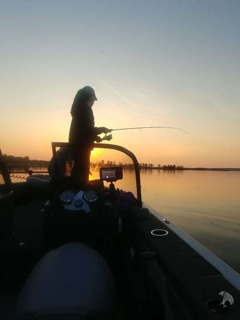 A young girl stands at the front of a motor boat, holding a rod, while the sun rises in the background.