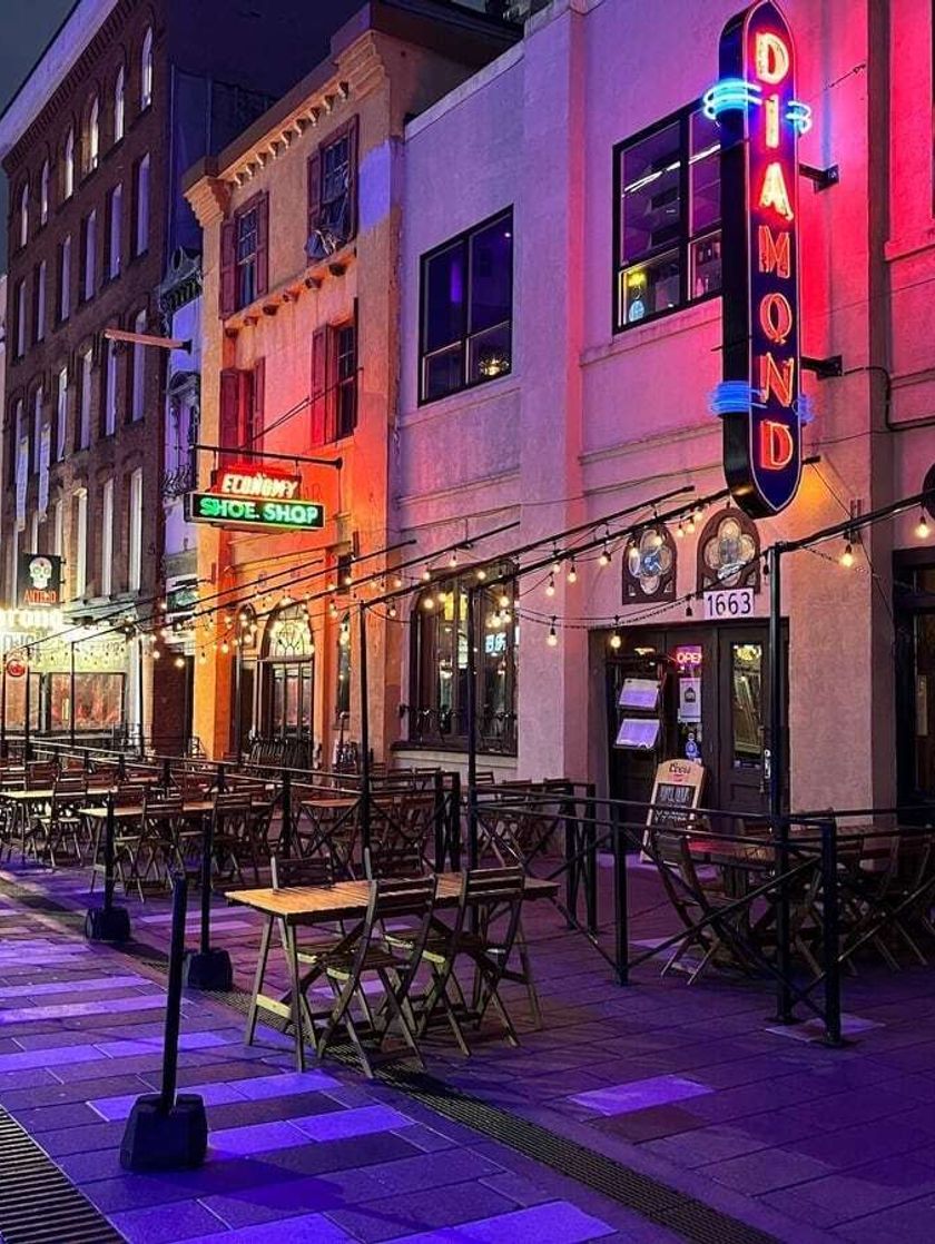 A cobbled streetscape at night, with dining tables and strings of lights set out in front of a row of midrise buildings. A vertical neon sign in the foreground says Diamond.
