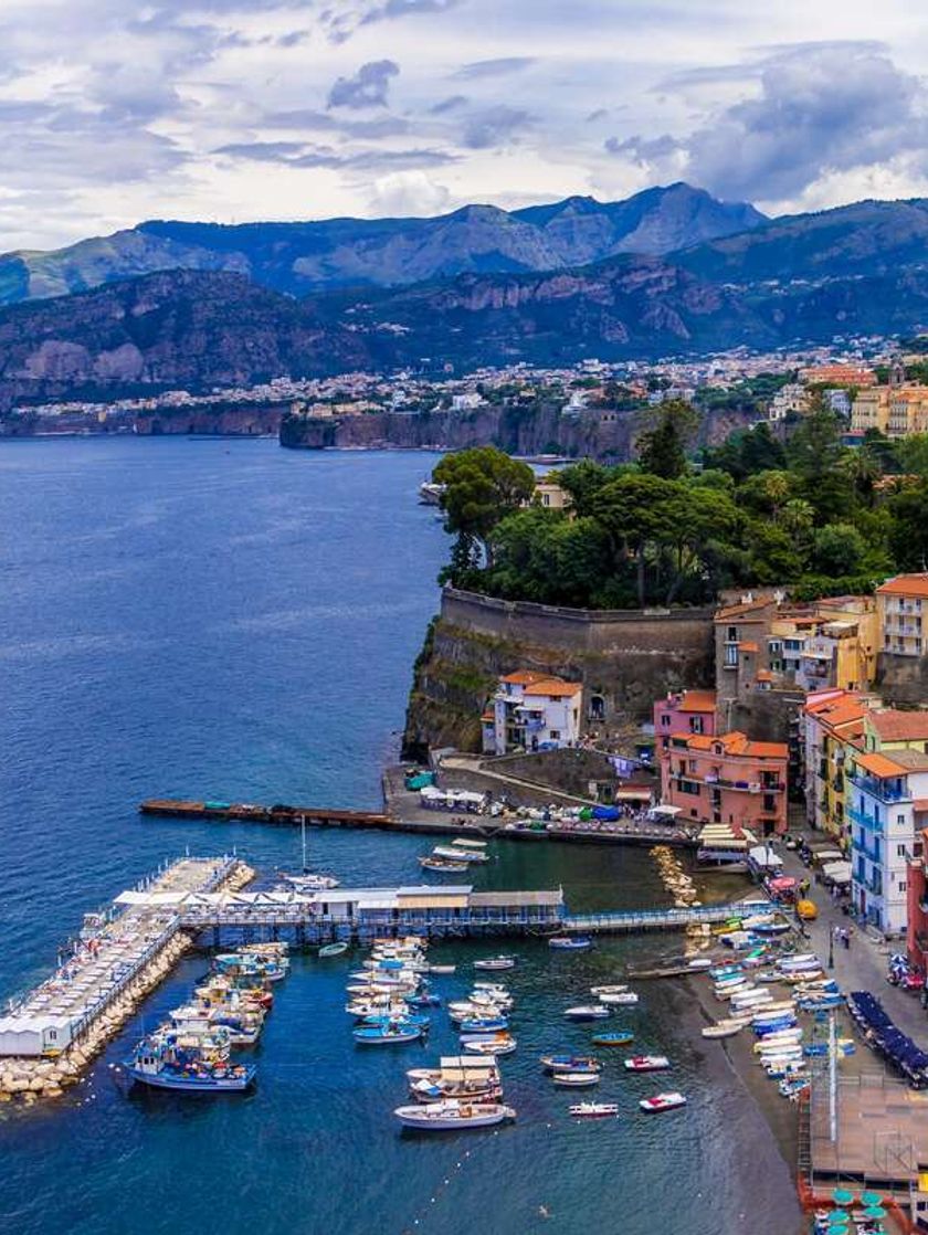 The coastline of Sorrento with a marina and boats in the bay, orange and pink low-rise buildings dotting the landscape, and rocky cliffs in the distance.