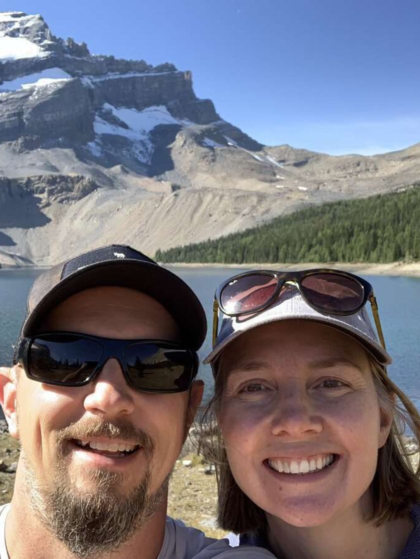 A selfie of a man and woman wearing baseball caps and sunglasses with a view of a lake, pine trees and snow-capped peaks behind them.
