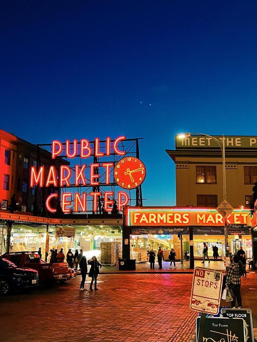 A pink-ish red neon sign with a large clock and lettering that says Public Market Area glows over a cobbled area where people walk. Another neon sign says, Farmers market. 